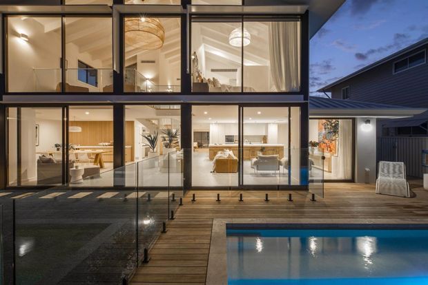 Twilight exterior view of a contemporary multi-storey residence with extensive floor-to-ceiling glazing, dark framing and cladding, a timber pool deck, and a swimming pool in the foreground. Warmly illuminated interiors reveal open-plan living, dining and kitchen areas, with an upper-level bedroom or living space visible through the glazing.