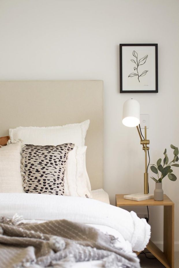 A softly styled bedroom corner with a cream upholstered bedhead, layered white bedding, a patterned accent cushion, and a timber bedside table. A slim brass and white floor lamp, small potted plant, and framed botanical artwork add warm decorative detail against a light neutral wall.