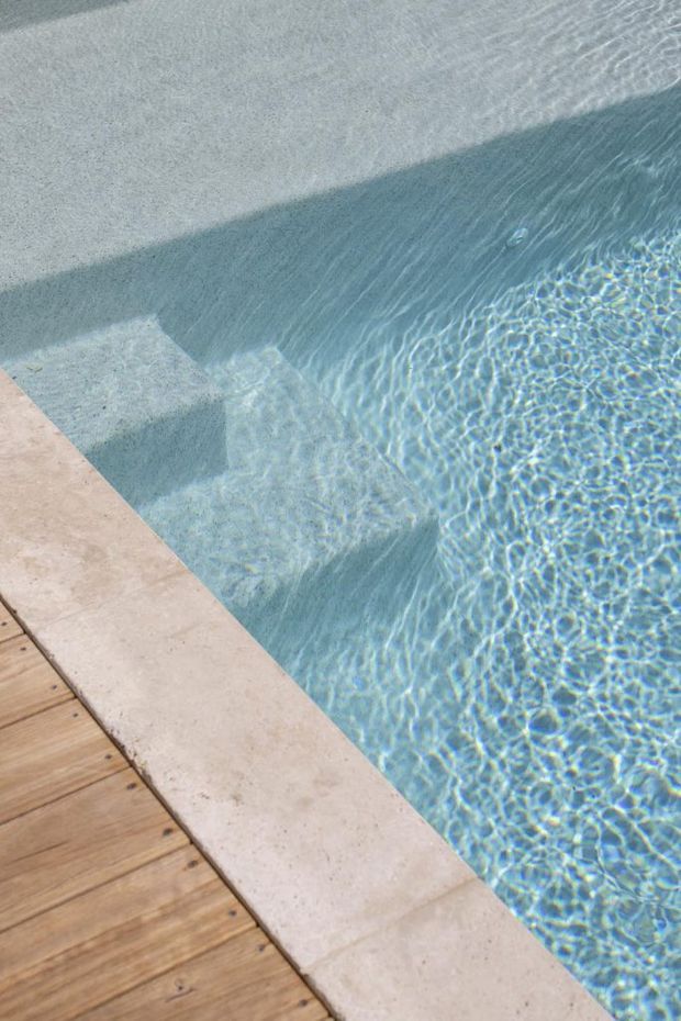 Close-up view of a clear swimming pool with visible submerged steps, pale stone coping, and adjacent warm timber decking. Sunlight creates rippling reflections across the blue water.