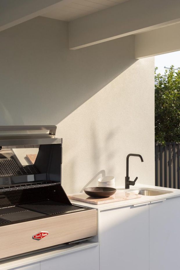 A compact outdoor kitchen or barbecue area with a built-in stainless-steel and timber-finished barbecue, white cabinetry, pale stone benchtop, integrated sink, and matte black gooseneck tap. The space is sheltered by a roof with a white-painted ceiling and opens to a fenced garden or outdoor area, with strong sunlight creating angular shadows on the rendered wall.