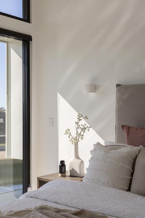 A softly lit contemporary bedroom with a neutral palette, large black-framed glazing, a tall upholstered bedhead, layered white bedding, and a simple timber bedside table styled with a sculptural vase and small dark bottle. Strong sunlight creates geometric shadows across the white wall.