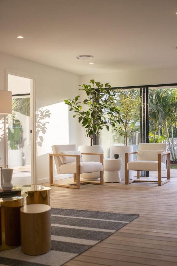 A bright, serene living room opening onto a timber deck, furnished with two light upholstered armchairs, a small sculptural side table, indoor trees, and a neutral striped rug. Full-height black-framed glazing connects the room to a leafy outdoor area, while recessed ceiling lighting and pale walls create an airy contemporary setting.