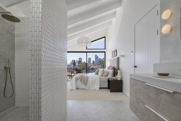Bright contemporary bedroom suite viewed from an adjoining bathroom. The foreground features a walk-in shower with small white vertical mosaic tiles and brushed metal fittings, while the bedroom beyond has a low-profile bed, soft neutral bedding, a dark bedside table, and large city-facing glazing. White-painted exposed ceiling beams, a pale floor, built-in cabinetry, and spherical wall lights create an airy, refined interior.