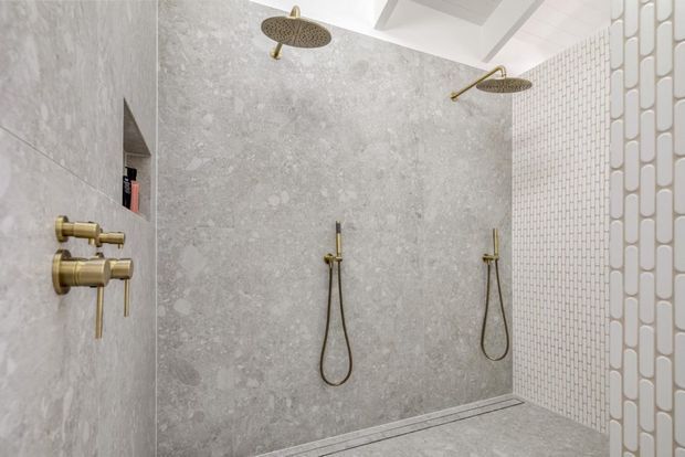 A bright contemporary walk-in shower with dual rainfall showerheads and handheld fixtures in brushed brass. Large-format grey stone-look wall tiles contrast with a white small-format mosaic-tile wall, while a partial glass screen and recessed niche complete the clean, refined bathroom design.