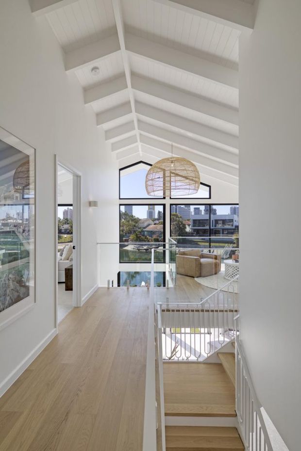 Bright upper-level hallway and stair landing in a refined coastal-style home, featuring a high pitched white-painted timber ceiling with exposed beams, pale timber flooring, white walls, a glass stair balustrade, and large black-framed windows overlooking an urban waterfront setting. A woven pendant light and adjacent living area are visible beyond the landing.