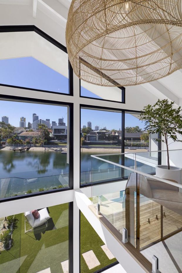 A bright, contemporary double-height interior overlooking a calm waterfront canal and urban skyline. Floor-to-ceiling black-framed glazing forms a dramatic window wall, with a glass balustrade around an upper-level landing or balcony. A large woven rattan pendant light hangs overhead, while a potted indoor tree and neutral upholstered seating soften the space. A landscaped lower courtyard with artificial grass and outdoor seating is visible below.