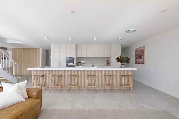 Bright open-plan kitchen and living area with a long white island bench, six timber bar stools, white handleless cabinetry, pale stone-look flooring, and a light neutral palette. A timber-lined stair enclosure and partial tan sofa are visible at the left, with artwork on the right wall.