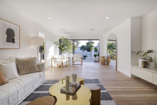 Bright, airy contemporary living room with a white slipcovered sofa, sculptural coffee table, patterned area rug, pale timber flooring, built-in white cabinetry, indoor plants, and full-width glazing opening toward an outdoor terrace. The room has a calm neutral palette with warm wood accents and abundant natural light.