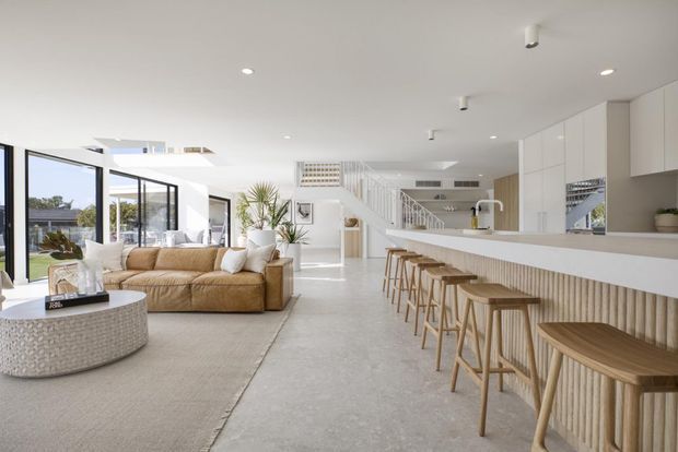 Bright open-plan contemporary interior combining a living room and kitchen. The space features a large beige sectional sofa, textured round coffee table, expansive glazing, a long white kitchen island with integrated seating, light timber stools, white cabinetry, and a staircase leading toward the rear of the home. Pale polished concrete flooring and abundant natural light create an airy, refined atmosphere.