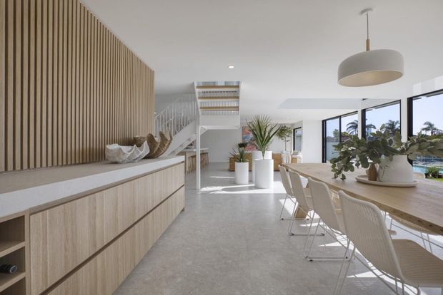 Bright, open-plan contemporary interior combining a kitchen and dining area. The space features pale timber cabinetry, a full-height vertical timber-slat feature wall, a long timber dining table with light upholstered wire-frame chairs, indoor plants, white pendant lighting, polished concrete-style flooring, and expansive black-framed glazing overlooking greenery and a pool.