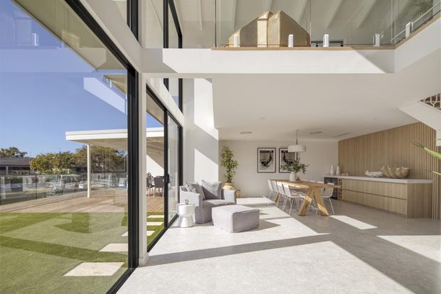 Bright contemporary double-height open-plan living and dining area with a built-in kitchen/bar zone, extensive floor-to-ceiling black-framed glazing, and a mezzanine or upper landing overlooking the room. Pale flooring, white walls, light timber cabinetry and vertical timber wall panelling create a clean, airy interior. The glazing opens toward a landscaped lawn and poolside or recreation area.