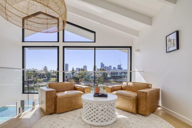 Bright contemporary living room with vaulted exposed-beam ceiling, expansive black-framed glazing, and a glass balustrade overlooking a waterfront urban view. Two tan leather armchairs and a matching sofa surround a round white lattice coffee table, with a large woven pendant light and pale patterned rug adding texture.