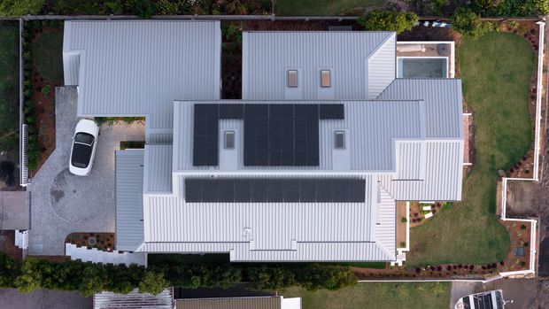 Aerial top-down view of a contemporary residential property with multiple interconnected pitched roof volumes, extensive grey metal roofing, and large dark solar panel arrays. The home is surrounded by landscaped gardens, paved outdoor areas, fencing, and a driveway with parked vehicles.