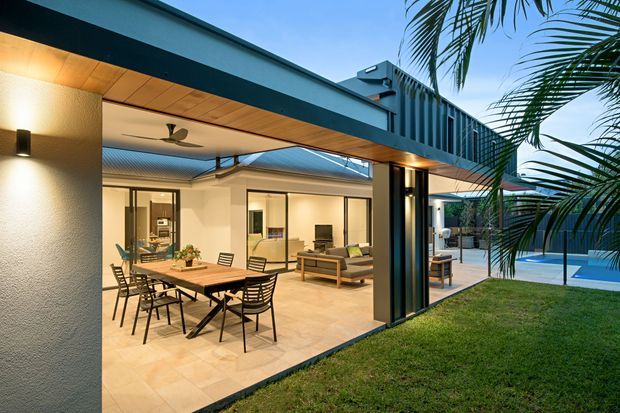 Contemporary single-storey home photographed at dusk, featuring a broad covered outdoor dining and living area opening through extensive floor-to-ceiling glazing toward a landscaped yard and swimming pool. The architecture combines a flat projecting roof, dark metal structural framing, textured light walls, warm timber soffit lining, and open-plan indoor-outdoor living. A dining table and chairs occupy the covered terrace, while a sofa and coffee table are visible inside.