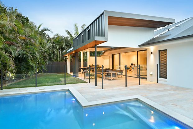 Contemporary two-storey residential exterior viewed from a landscaped pool area. The home features crisp white walls, dark vertical metal cladding, projecting flat-roofed upper volumes, extensive glazed openings, slim dark frames, and a covered outdoor entertaining area opening onto a pale tiled terrace. A swimming pool and tropical planting occupy the foreground and side garden.