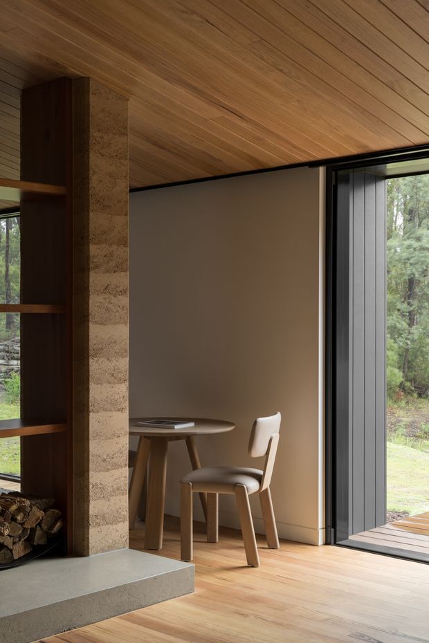 A warm, minimalist interior corner featuring a compact wooden writing desk and chair beside a substantial textured masonry fireplace with built-in timber shelving and stacked firewood. Full-height dark-framed glazing opens to a wooded landscape, while timber boards cover the ceiling and floor.