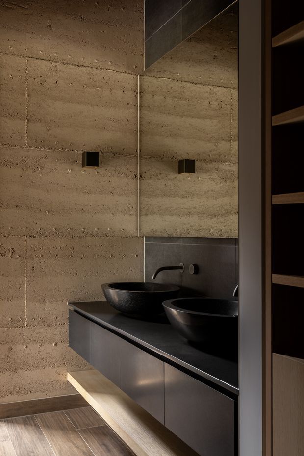 A moody contemporary bathroom vanity with two dark vessel basins, black wall-mounted faucets, and a large mirror. The walls are formed from rough exposed concrete blocks, with dark cabinetry, a dark stone-like benchtop, and warm timber open shelving and drawer detailing. Timber-look flooring and restrained lighting create a refined, industrial atmosphere.