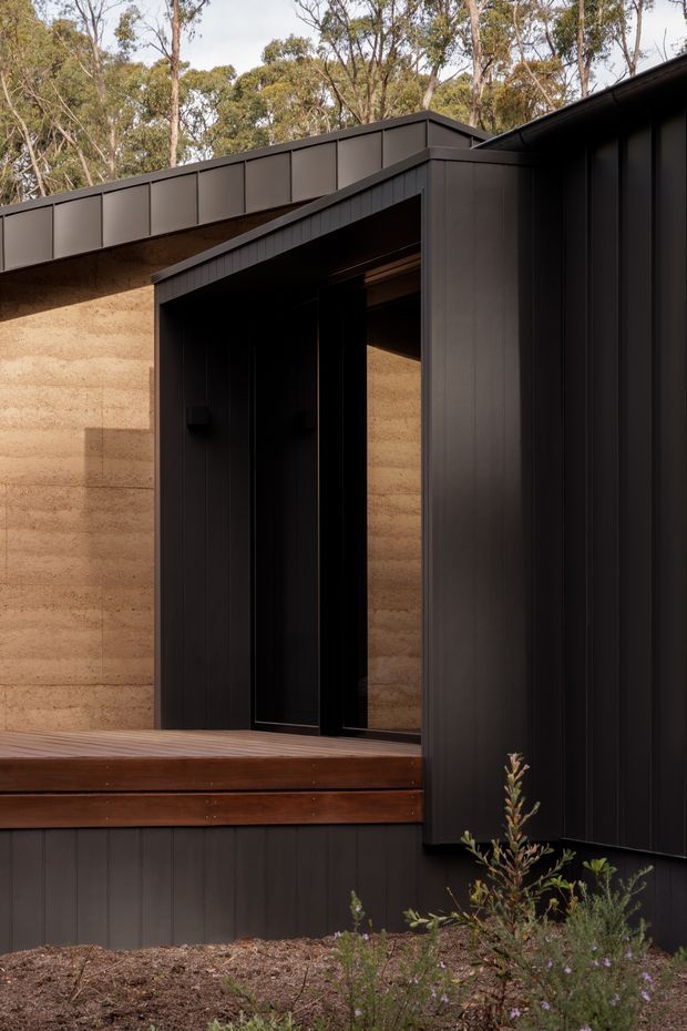 Close-up exterior view of a contemporary dark-clad building set among eucalyptus trees. The facade combines vertical dark metal cladding with warm textured earthen walls, a recessed black entry, and a raised timber deck. The angular roof and entry canopy create a strong geometric composition, with native planting in the foreground.