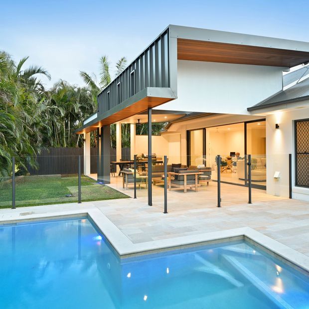 Exterior view of a contemporary multi-level home with dark vertical cladding, white rendered walls, and warm timber-lined soffits. The covered alfresco living and dining area opens onto a large pale-paved terrace beside a swimming pool, with glass balustrades and lush tropical planting surrounding the property.