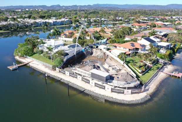 Aerial view of a waterfront residential construction site on a narrow peninsula, surrounded by canals and established suburban homes. The site includes retaining walls, construction fencing, earthworks and partially built structures, with mountain ranges visible in the distance.