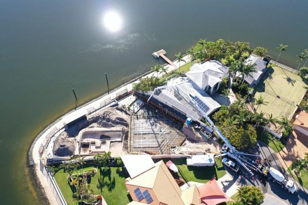 Aerial view of a waterfront residential property with a curved retaining wall and shoreline, landscaped gardens, several light-coloured pitched-roof buildings, tennis court, dock, and an active construction area with structural framing and construction equipment.