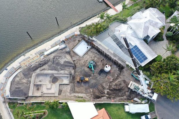 Aerial view of a waterfront residential property under construction, showing excavation, concrete foundation work, construction machinery, temporary site fencing, and a large house with light-coloured roofs and rooftop solar panels. A shoreline and landscaped gardens are visible around the site.