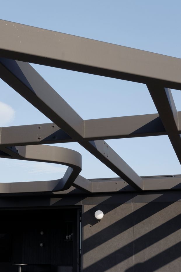 Close-up exterior architectural view showing a dark contemporary building facade beneath an exposed geometric steel pergola or roof structure. Large rectangular and diagonal beams form a strong angular pattern against a clear blue sky, casting dramatic shadows across the wall. A curved steel beam and a small round exterior wall light are visible.