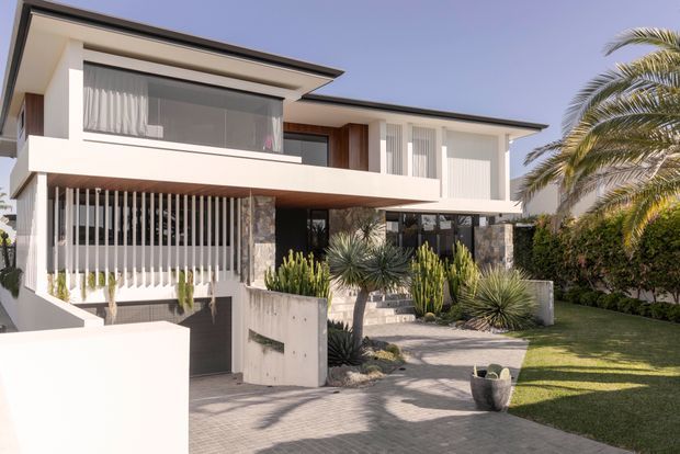 Contemporary two-storey residence with a flat roof, crisp white rendered walls, extensive glazing, timber soffits, vertical privacy screens, and natural stone-clad columns. The front entry is approached by a paved path through sculptural drought-tolerant landscaping, with lawn and palm planting to the side.