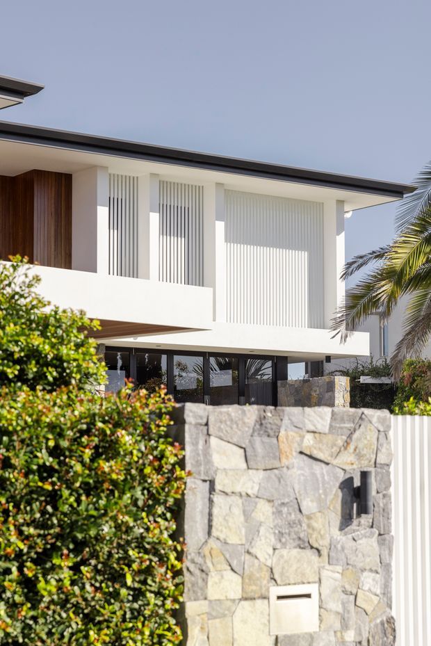 Contemporary two-storey residential facade with a low-pitched dark roof, crisp white rendered walls, vertical white screening and a warm timber-clad entry panel. Large glazed openings sit beneath projecting upper-level forms. The front boundary features a substantial irregular natural-stone wall with an integrated mailbox, alongside manicured hedging and a palm tree.