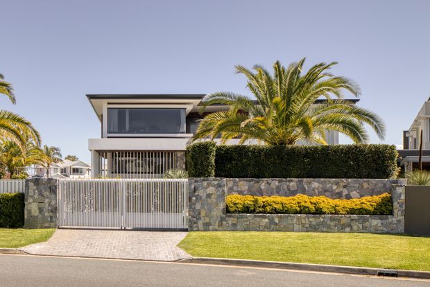 Street-facing view of a modern two-storey residence with a projecting flat roof, dark-framed glazing, vertical metal security screens, and a tall sliding vehicle gate. The front boundary combines irregular grey stonework, clipped hedging, and a raised planter with bright yellow-green foliage, opening onto a neatly paved driveway and lawn. Palm trees and neighbouring homes reinforce a warm suburban setting.