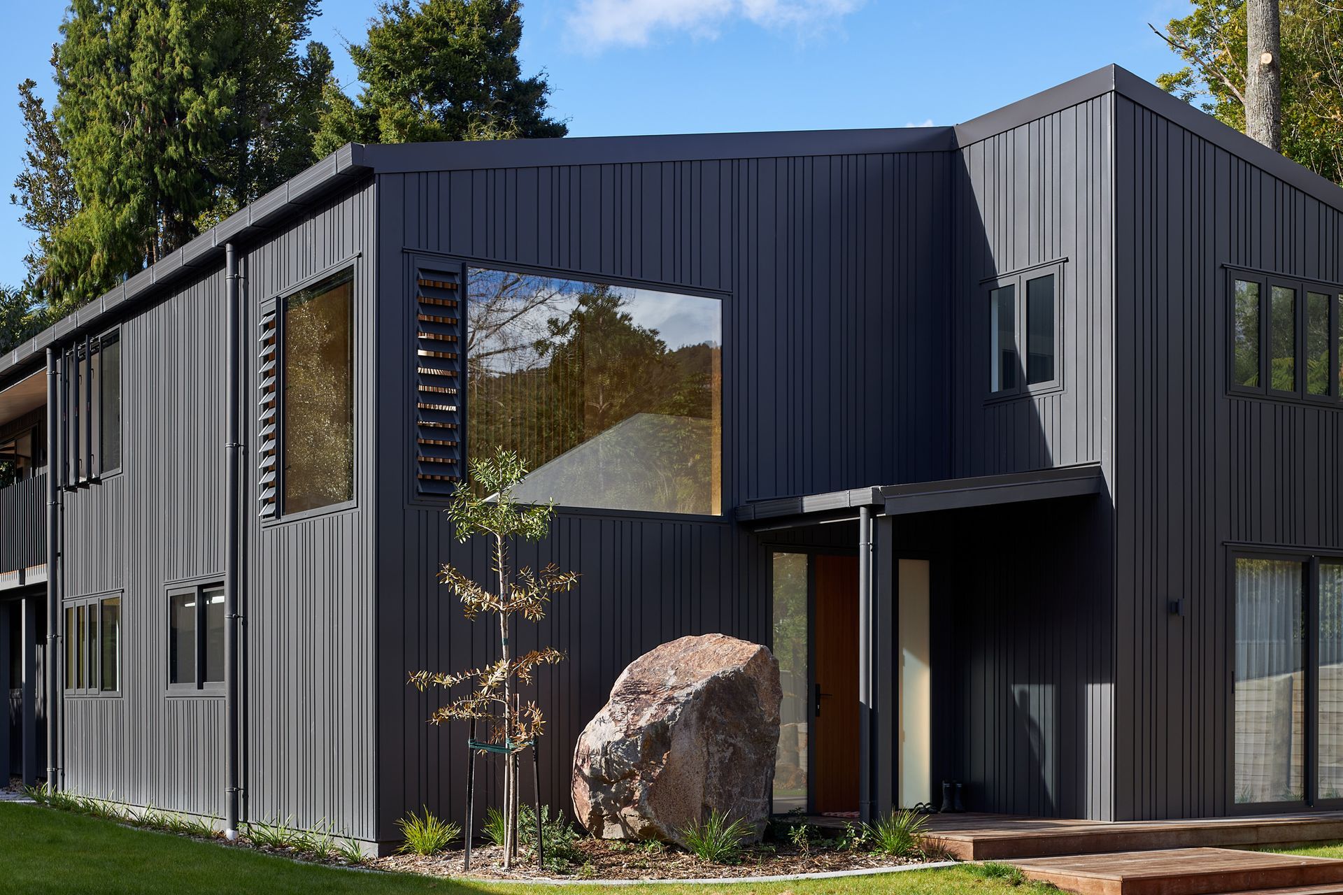 The entrance to The Owl's Retreat is defined by a restrained palette of slate grey Nu-Wall cladding and a sculptural boulder that anchors the arrival sequence. Large areas of glazing reveal the warmth of the plywood-lined interior, creating a welcoming contrast between the robust exterior and the natural materials within.