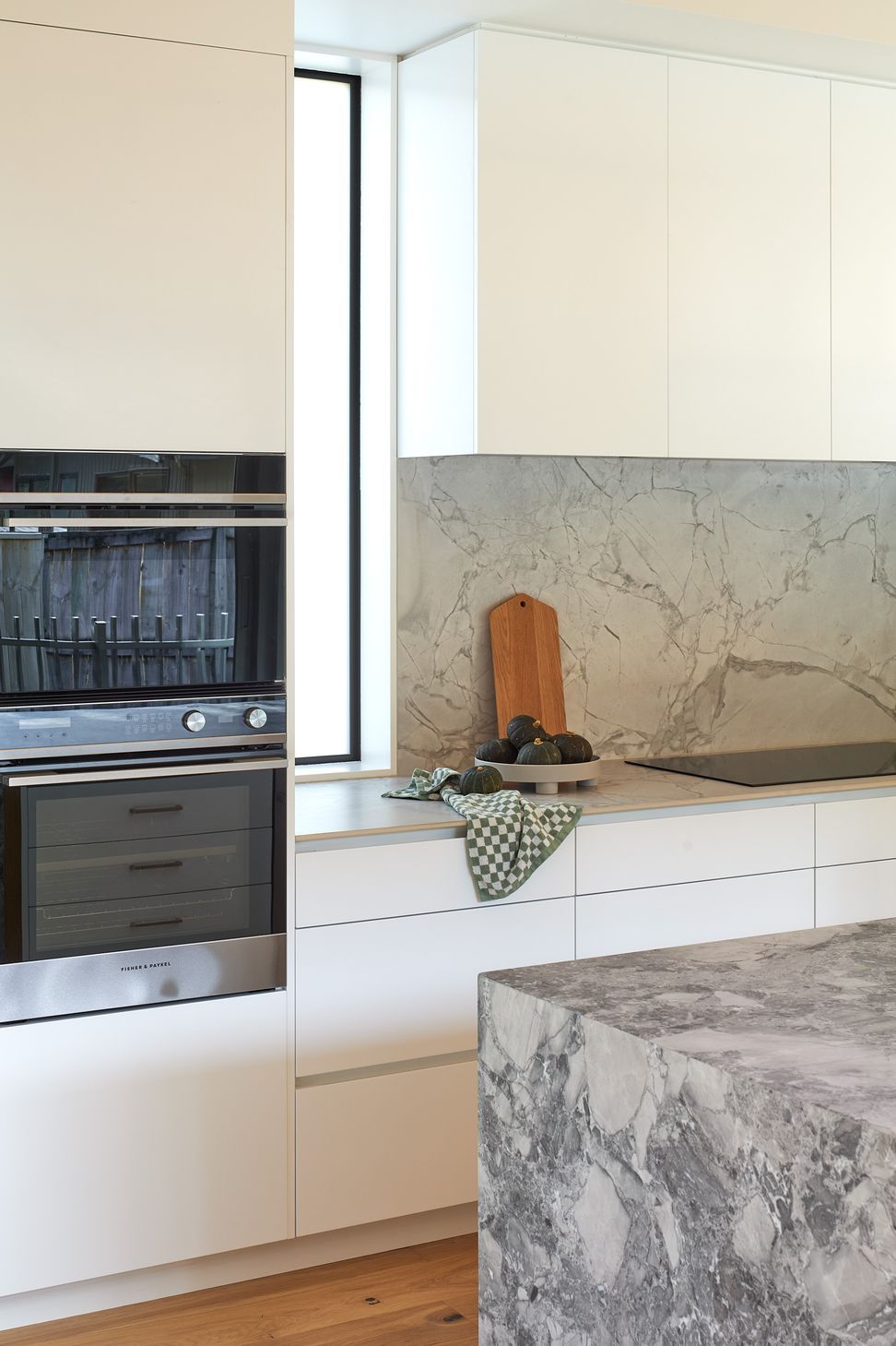 Designed to bring people together, the kitchen balances clean, contemporary detailing with the warmth of natural materials. Crisp white cabinetry is complemented by Super White granite benchtops, while a porcelain work surface and splash-back create a seamless, durable backdrop for everyday use. Overhead, birch plywood ceilings introduce warmth and texture, giving the space a relaxed, inviting character that connects effortlessly with the adjoining dining and outdoor living areas.