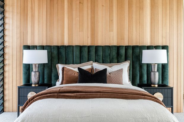 A warmly styled bedroom with a bed centered against a full-height vertical timber panel wall. The bed features a deep green vertically channelled upholstered headboard, layered white and brown bedding, and assorted cushions. Matching dark bedside tables with ceramic lamps flank the bed, while a dark louvred screen is visible at the left edge.