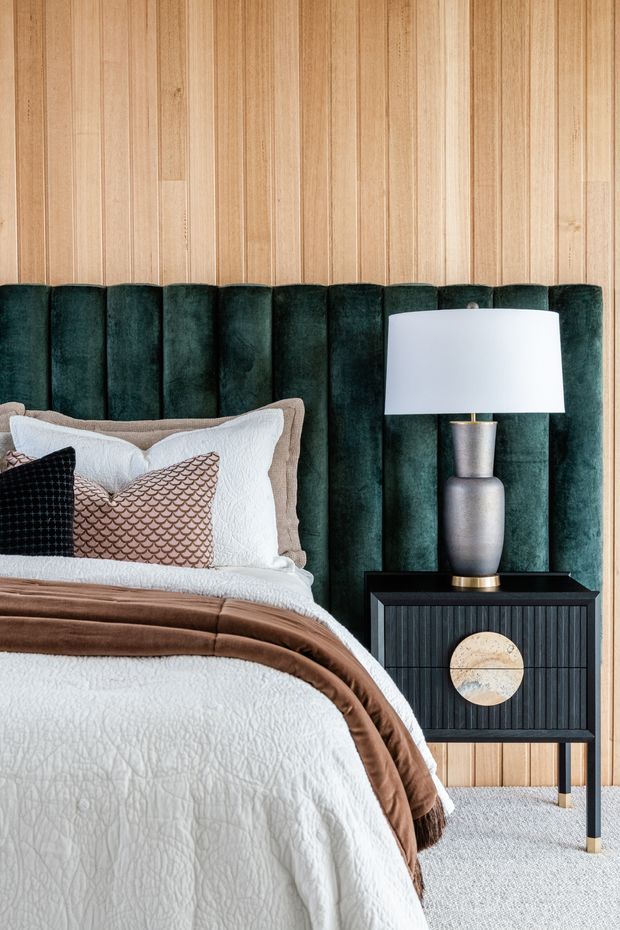 A close-up view of a styled bedroom featuring a bed with layered white bedding, a brown throw, and decorative cushions. The bed has a tall, channel-tufted dark green upholstered headboard set against a vertical light timber-panelled wall. A black fluted bedside table with a round decorative inlay supports a metallic lamp with a white drum shade. Light carpet covers the floor.