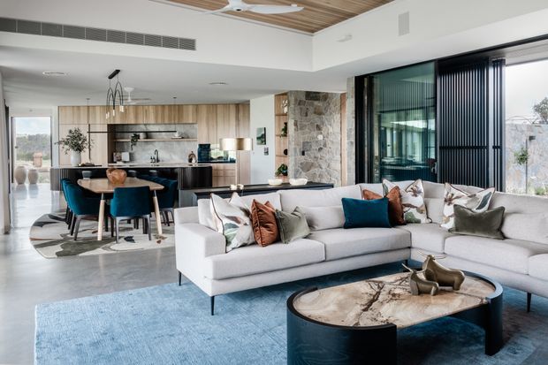 A bright, open-plan contemporary living space with a large sectional sofa, blue area rug, sculptural round coffee table, and expansive dark-framed glazing with vertical blinds. The living room opens to a dining area and kitchen, with warm timber ceiling lining, pale timber cabinetry, a stone-clad fireplace or feature wall, and polished concrete flooring.