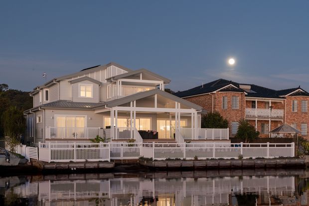 Exterior dusk view of a large multi-storey waterfront residence with light weatherboard cladding, multiple gabled roof forms, extensive covered verandas and balconies, white railings, and a fenced edge beside calm reflective water. A neighbouring brick home is visible to the right.