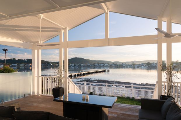 A bright covered waterfront outdoor living area with a white-framed, open-sided structure, pitched translucent roof, timber decking, outdoor seating, and a central table. The space overlooks calm water, a pier, and a settled shoreline with low hills beyond.