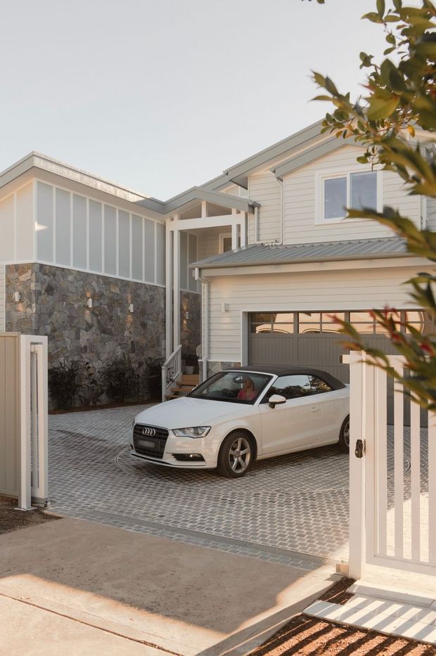 Exterior view of a contemporary multi-level residence with pale horizontal weatherboard cladding, a grey metal gabled roof, and a prominent stone-clad wall. The property includes a covered entry, garage with a glazed-panel door, front steps, a metal gate, and a paved driveway. A car is present in the driveway but is not part of the architectural classification.