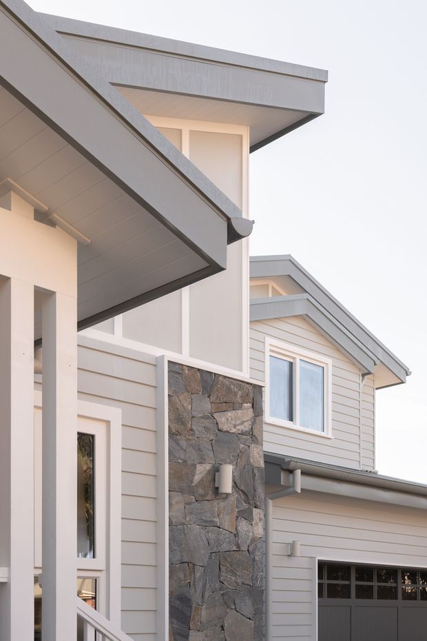 Close-up exterior view of a contemporary multi-level home featuring pale horizontal weatherboard cladding, angular projecting roof forms, dark trim, a natural rough-stone feature wall, tall glazing, and an integrated garage door.
