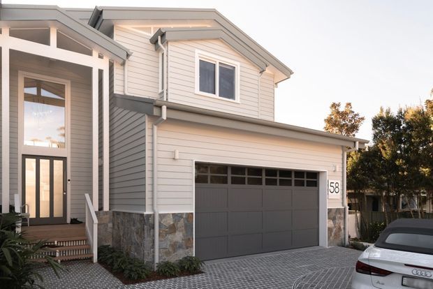 Exterior view of a contemporary multi-level residential home with light horizontal weatherboard cladding, grey trim, a large integrated double garage, tall glazed entry, and a stone-clad lower section. The driveway is paved in a diagonal grid pattern, with mature trees and a parked car at the edge of the frame.