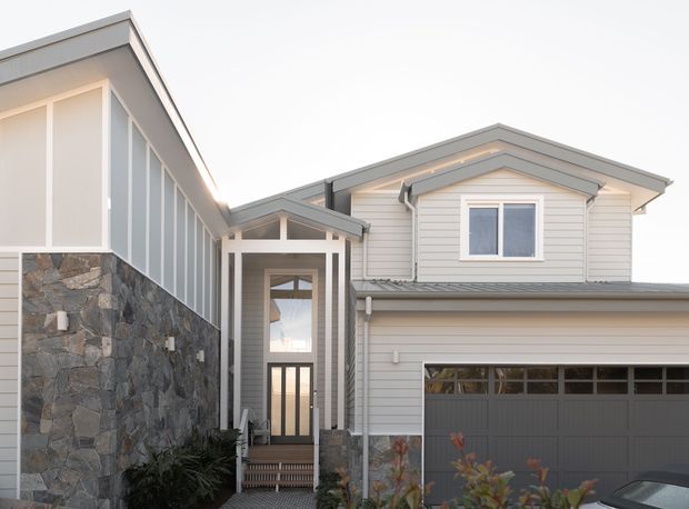 Exterior view of a contemporary multi-level residence with light grey horizontal weatherboard cladding, a dark grey garage door, large glazed entry, and a prominent natural stone-clad wall. The house has multiple pitched roof forms and an attached garage, with a narrow landscaped approach in the foreground.