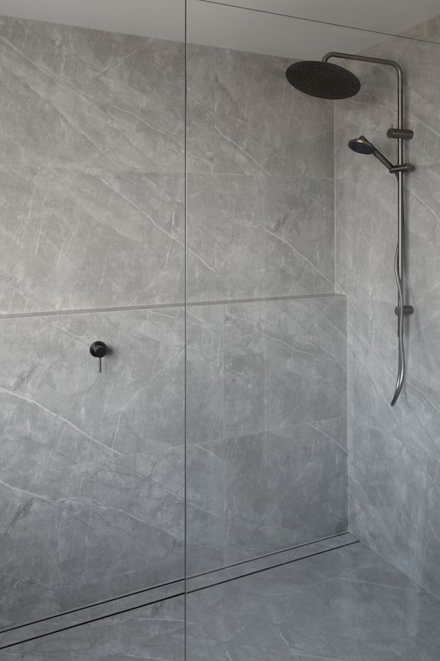 Minimalist walk-in shower enclosed by a clear frameless glass screen. The walls and floor are clad in large-format grey marble-look tiles with subtle veining, while matte dark metal fixtures include a rain shower, handheld shower, controls and linear floor drain.