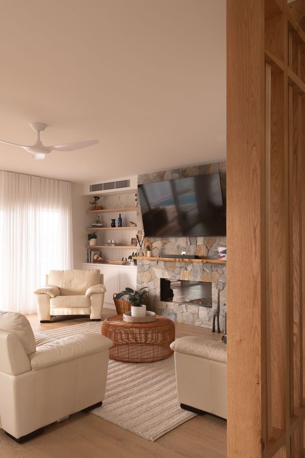 Warm, sunlit living room viewed through a timber-framed opening. The room features cream leather seating, a woven round coffee table, a neutral textured rug, sheer curtains, built-in shelving, and a prominent rough-stone fireplace wall with a wall-mounted television. Natural timber framing and joinery add a relaxed, earthy character.
