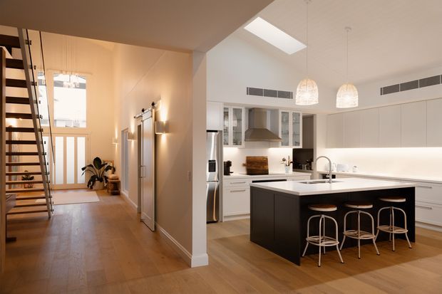 Open-plan contemporary home interior showing a bright white kitchen with a dark island and bar stools, adjoining a timber-floored entry or hallway with a floating staircase, skylight, pendant lights, and warm wall lighting. The kitchen features white cabinetry, integrated appliances, a tiled or light benchtop, and a sculptural rangehood.
