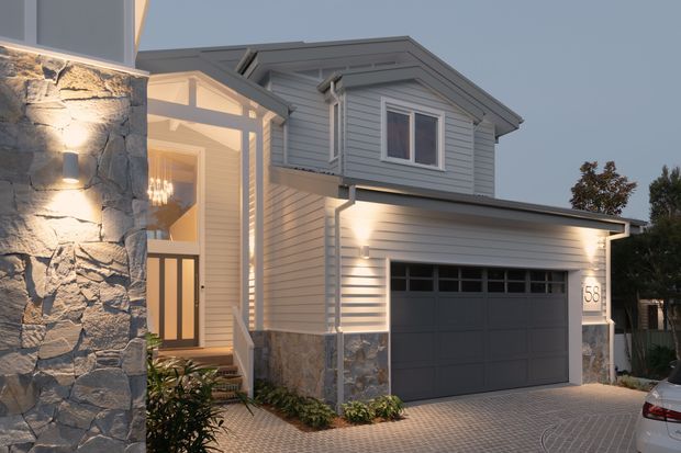 Twilight exterior view of a two-storey residential home with a prominent double garage, grey horizontal weatherboard cladding, stone veneer accents, a gabled roof, and a glazed entry. The facade is illuminated with warm architectural lighting, and the driveway is paved in a repeating pattern.