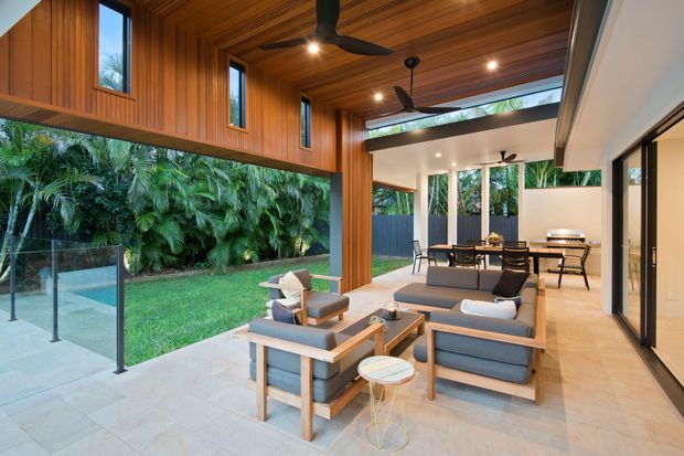 A spacious, open-plan indoor-outdoor living area with a covered patio, lounge seating, dining area and kitchen visible beyond. The space features a dramatic timber-lined ceiling and upper wall, extensive glazing, slim dark window and door frames, ceiling fans, pale stone or tiled flooring, and a lush planted courtyard garden enclosed by frameless glass balustrades.