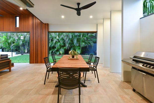 A bright, open-plan dining and outdoor entertaining area with a long timber dining table and black chairs, pale tiled flooring, warm vertical timber wall cladding, large openings to lush greenery, a sculptural ceiling fan, and a built-in stainless-steel barbecue along the right wall.