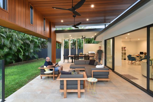 A spacious covered outdoor living and entertaining area connected to a contemporary home. The space features a timber-lined ceiling with ceiling fans, large-format pale stone or concrete-look paving, timber-framed dark upholstered lounge furniture, a dining setting, and extensive black-framed sliding glass doors opening to a bright interior. Lush tropical planting and a lawn border the courtyard, creating a warm indoor-outdoor connection.