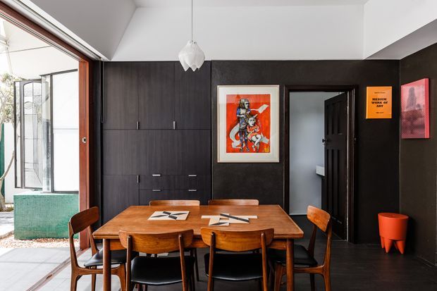 A compact, dark-toned dining area with a solid timber dining table and mid-century-style wooden chairs. Full-height dark built-in cabinetry lines the rear wall, alongside framed artwork, a black internal doorway and a small orange side stool. White ceiling planes and a sculptural pendant light provide contrast, while glazed doors at left open toward an outdoor area.