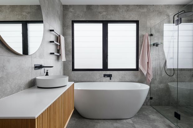 Contemporary bathroom with a freestanding white bathtub, wall-mounted round basin, fluted timber vanity cabinetry, large grey stone-look wall and floor tiles, black-framed windows, and a frameless glass shower enclosure. The space has a clean, monochromatic palette with warm timber detailing.